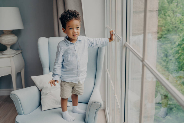 Teljes hossza kíváncsi kis afro amerikai fiú édes kisgyerek elegáns kopás néz ki az ablakon, és várja anya, miközben töltik szabadidő és játszik otthon a fény szobában - Fotó, kép