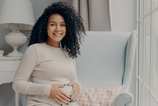 Portrait de jeune femme afro-américaine enceinte souriante en tenue décontractée se relaxant après avoir marché dehors sur un fauteuil dans le salon, en gardant les mains sur le ventre et en regardant la caméra avec une expression heureuse - Photo, image