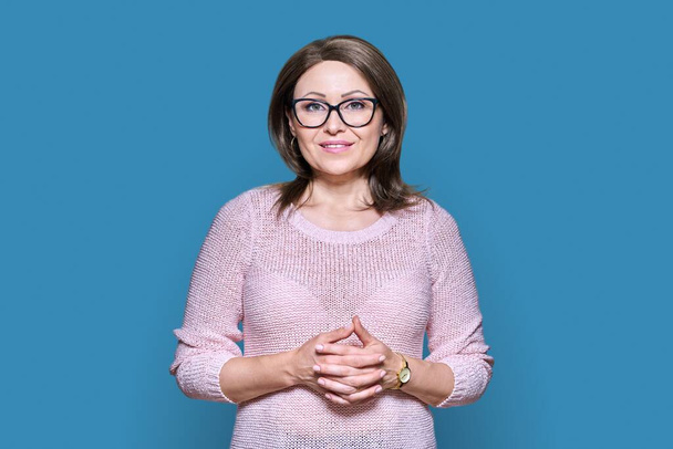 Attractive middle aged woman looking at camera on blue color background. Smiling 40s female with glasses, folded hands. Lifestyle, confidence, calmness, mature people concept - Zdjęcie, obraz