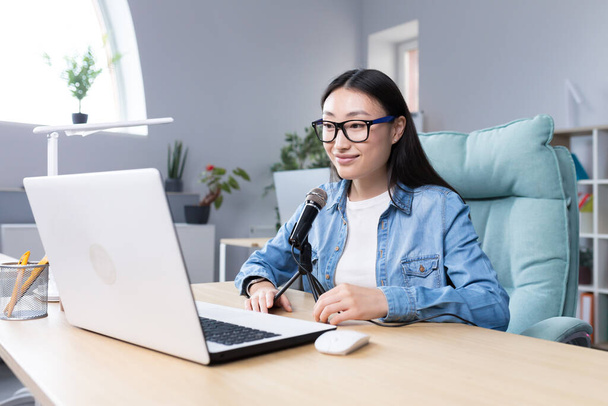 Podcast recording, interview. Young beautiful Asian woman sitting at a table with a microphone and a laptop. Talks, tells. Makes an audio recording of a podcast, gives an interview via video call. - Photo, Image