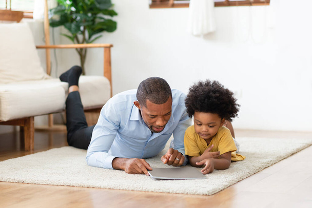 Black African American father and little son lying on floor and reading a book together at home. - Foto, Bild