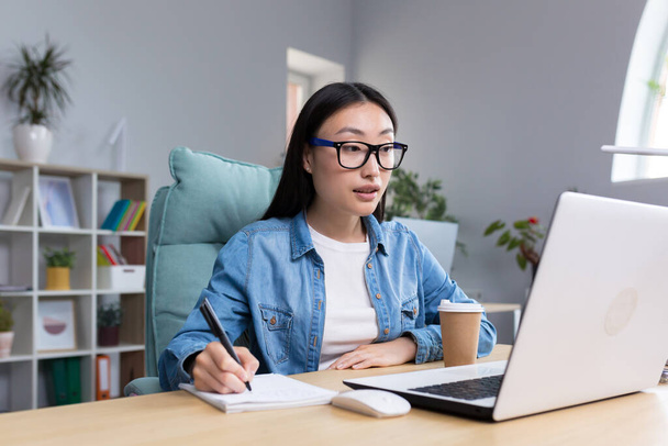 Distance Learning. Young beautiful Asian female student taking an online exam. Sitting in glasses and jeans at the table at the computer, making notes in a notebook. - Φωτογραφία, εικόνα