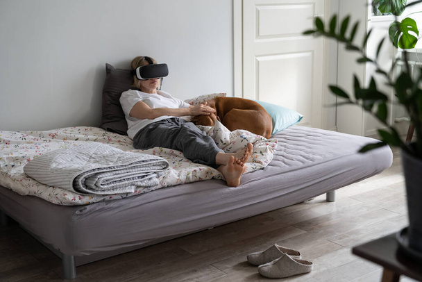 Single middle-aged woman spends weekend time playing VR games wearing VR glasses lying on bed. Lonely relaxed female in nightwear has playing games addiction staying home Dog only friend sleeps nearby - Zdjęcie, obraz