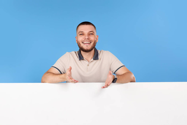 Portrait of cheerful bearded man leaning on empty placard for advertisement with empty copy space on blue background - Foto, Imagem