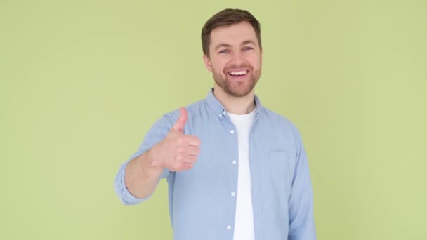 Young handsome man with beard in denim shirt on yellow-green background, sign of success making positive hand gesture, smiling and happy thumb up. Cheerful facial expression - Video