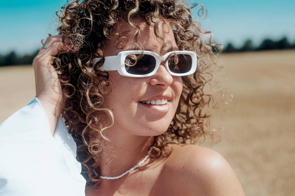 portrait of young curly haired woman in white dress with a white sunglasses posing in the wheat field - Fotó, kép