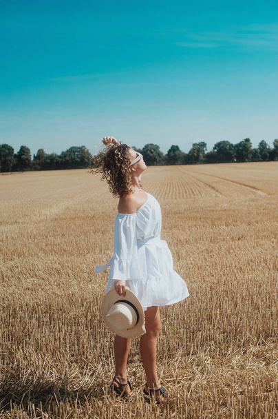 portrait of young curly haired woman in white dress with a white sunglasses posing in the wheat field - Foto, immagini