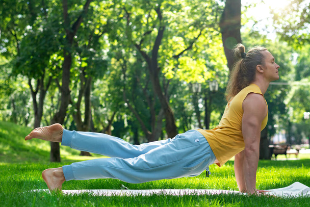 Young man practice yoga in the park. Yoga asanas in city park, sunny day. Concept of meditation, wellbeing and healthy lifestyle - Photo, image