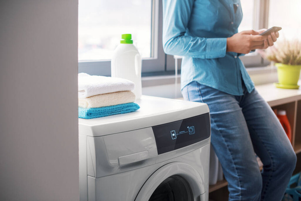 Woman doing laundry at home using a smart washing machine, she is controlling the smart washer from her smartphone - Foto, Imagen