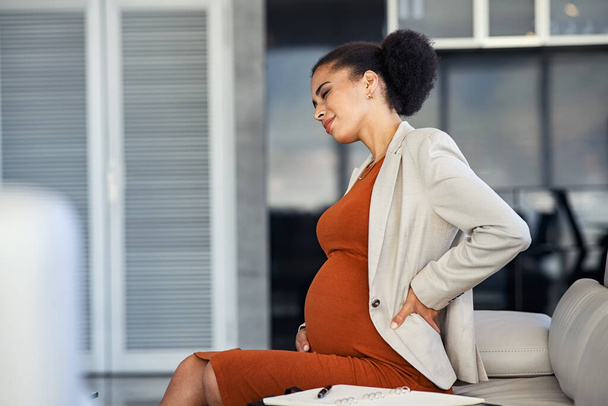 Young pregnant woman sitting on couch suffering from pain while working in office. Pregnant young woman feeling sick at office on couch with eyes closed. Black businesswoman suffering from back pain during pregnancy in modern office. - Photo, image