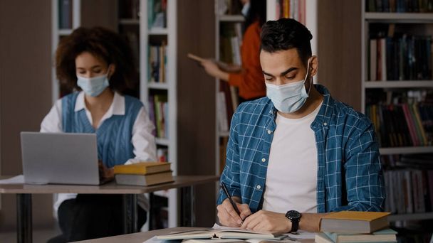 Öğrenciler kütüphanede çalışıyor koruyucu maskeli zeki Hintli çocuk masaya oturup ödevlerini yapıyor sınav hazırlıyor not defterine notlar yazıyor makale yazıyor. - Fotoğraf, Görsel