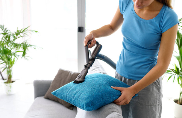 Young woman cleaning the couch in the living room, she is vacuuming the cushions using a vacuum cleaner - Foto, Bild