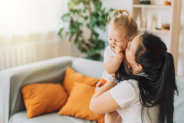 Young mother hugging and kissing her baby while sitting on the couch. - Valokuva, kuva
