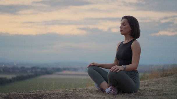 Полный вид на внимательную молодую женщину, делающую жест мудры, сидящую в позе лотоса на травянистом холме. Мирная девушка тысячелетия глубоко медитирует, делая дыхательные упражнения йоги в одиночку - Фото, изображение