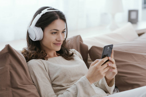 Young woman relaxing on the bed, she is wearing headphones and watching videos on her smartphone - Foto, Imagen
