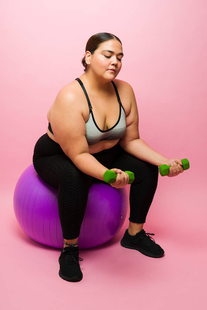 Big young woman using a fitness ball and lifting dumbbell weights while working out against a pink background - Zdjęcie, obraz