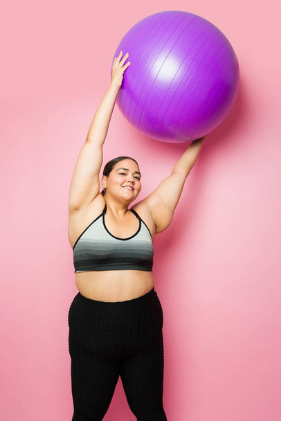 Портрет улыбающейся толстухи, поднимающей фитнес-мяч и занимающейся спортом во время активного образа жизни, чтобы похудеть - Фото, изображение