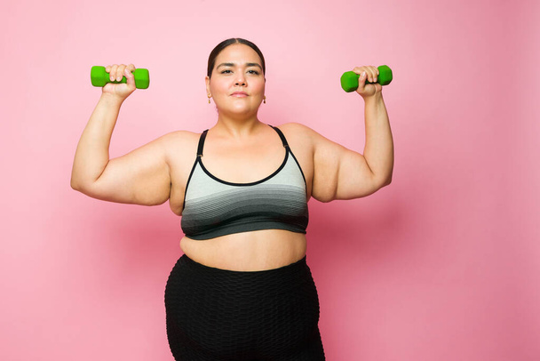 Obese hispanic woman looking at the camera while lifting dumbbell weights and exercising to lose some weight - Fotó, kép