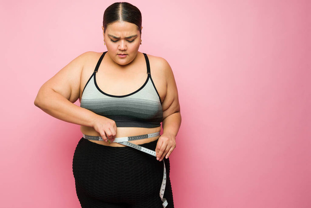 Worried overweight latin woman in activewear measuring her stomach after exercising to lose weight   - Фото, изображение