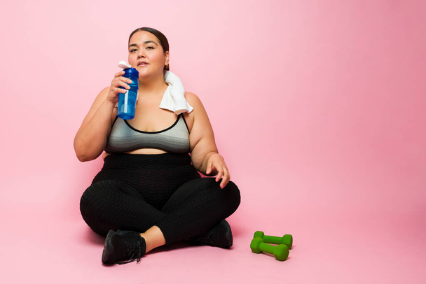 Активная женщина с избыточным весом с полотенцем для отдыха после тренировки и питья из бутылки с водой  - Фото, изображение