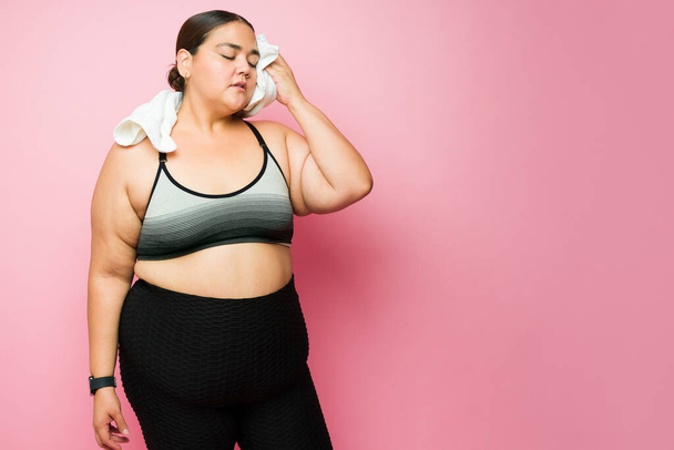 Exhausted plus size woman in sportswear sweating and looking tired after working out in front of a pink studio background - Fotó, kép