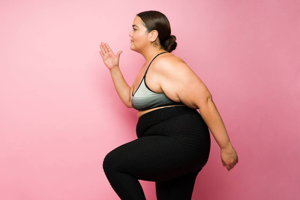 Side view of a sporty overweight woman running in place or jogging during her fitness workout against a pink background - Foto, imagen