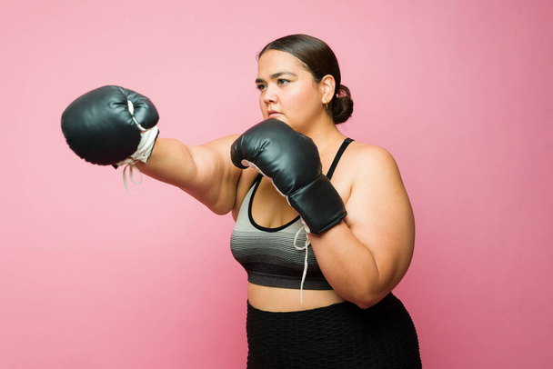 Overweight sporty young woman in activewear boxing using gloves and working out or training against a pink background - Photo, image