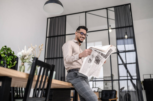 Newspaper, news. Smiling attractive well dressed man in glasses looking at newspaper standing leaning on table in modern apartment - Photo, image