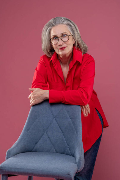 In a studio. Mature woman in red shirt and eyeglasses standing near the chair - Foto, Bild