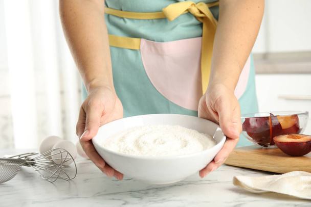 Woman holding bowl with flour at white marble table, closeup. Cooking of delicious plum cake - Fotó, kép