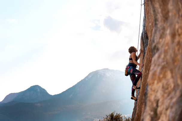 Una mujer trepa una roca contra un cielo azul, una chica fuerte entrena fuerza y resistencia, un deporte extremo, escalada en roca en terreno natural, un escalador sube con una cuerda - Foto, Imagen
