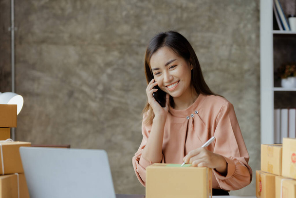 Asyalı bir kadın siparişi doğrulamak için bir müşteriyle telefonda konuşuyor, bir online mağazası var, özel bir nakliye şirketi aracılığıyla paketleyip gönderiyor. Çevrimiçi satış ve online alışveriş kavramları. - Fotoğraf, Görsel