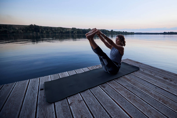 A fit yogi woman is practicing yoga on a dock at dusk. She is in the Boat yoga posture. - Foto, immagini