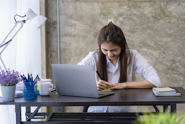 Посміхаючись, азіатська жінка працює з ноутбуком на своєму столі в сучасному офісі. Обчислення фінансових витрат робить записку на аркуші даних на графіку. - Фото, зображення