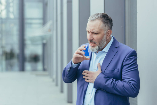 Senior gray-haired businessman sick has asthma attack difficult to breathe, man outside office building in business clothes uses inhaler to ease breathing - 写真・画像