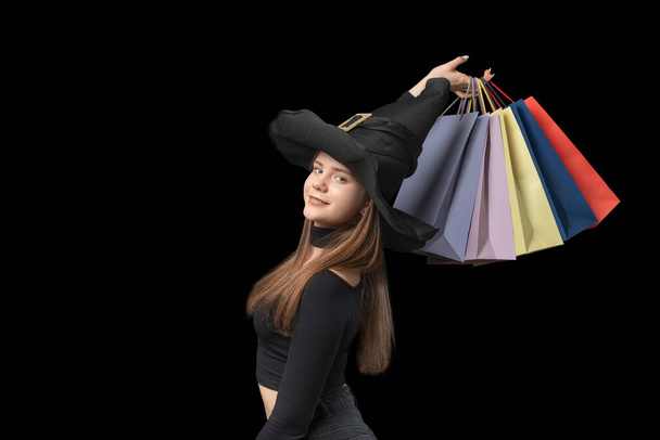 Portrait of girl in witchs hat with colorful paper shopping bags in hands. Black background. Copy space. Black Friday concept - 写真・画像