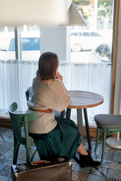 Young woman in long skirt sits at round table in cafe by window waiting for an order. Vertical frame - Foto, afbeelding