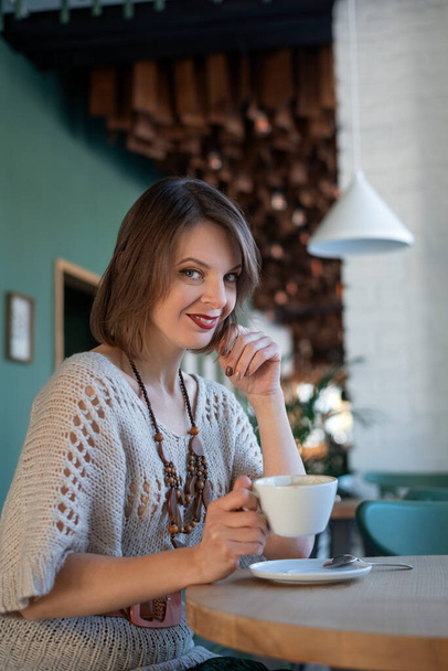 Smiling young woman in gray dress drinks coffee on inside cafe. Portrait of brunette girl sits at table in cafe - Photo, image