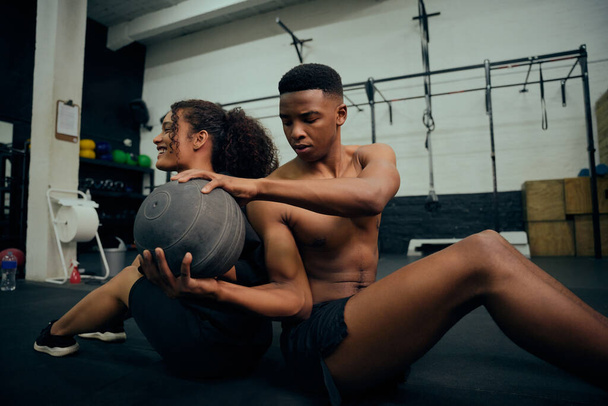 Multi-ethnic friends doing cross fit in the gym. African American male and female sitting down on the floor and passing a medicine ball during exercise routine. High quality photo. High quality photo - Φωτογραφία, εικόνα