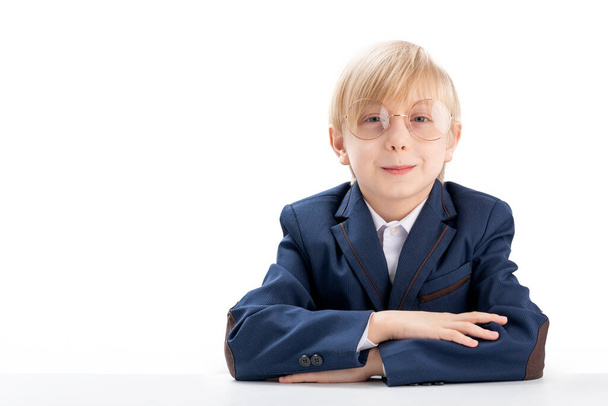 Elementary school student wears school uniform and glasses sit idly by at desk. Portrait of blond schoolboy on white background. Copy space - Foto, Imagen