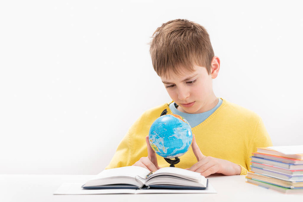 Boy in yellow jumper does geography lessons. Schoolboy sits at table and carefully studies the globe - Fotografie, Obrázek