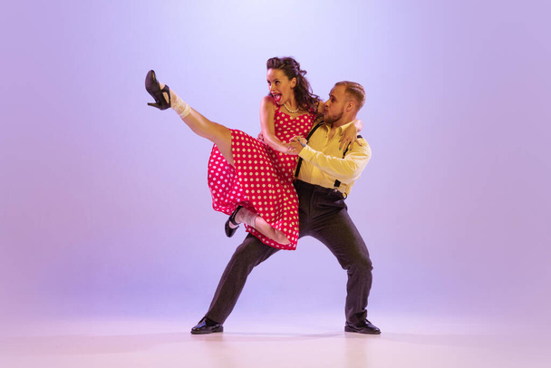 Lindy Hop. Active and emotional couple in colorful retro style costumes dancing incendiary dances isolated on purple background in neon. Actors in motion and action. Concept of art, 60s, 70s culture - 写真・画像