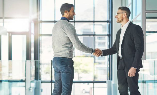 Businessman standing, smiling and with a handshake, greets a colleague in an office. Executive meets client or partner and shakes hands. Two happy professionals agree on a successful business project. - Foto, afbeelding