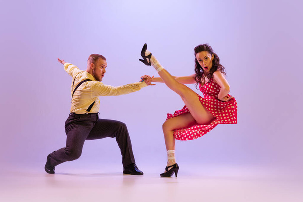 Beautiful girl and man in colorful retro style costumes dancing incendiary dances isolated on lilac color background in neon light. Actors in motion and action. Concept of art, 60s, 70s culture - Foto, imagen