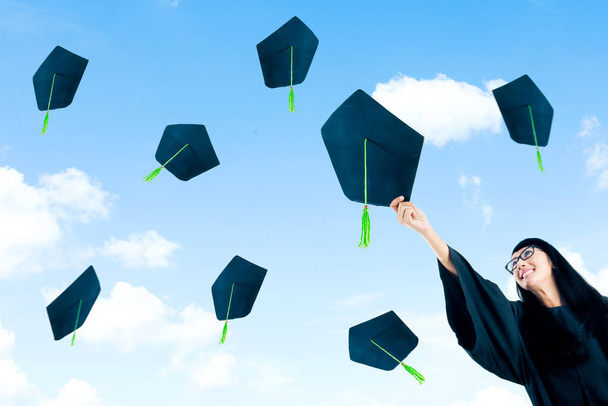Low angle view of happy female college student throwing a graduation hat while standing with blue sky background - Foto, afbeelding