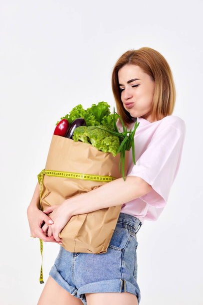 funny young woman holding paper bag with vegetables and measuring tape on white background. healthy food - Photo, image