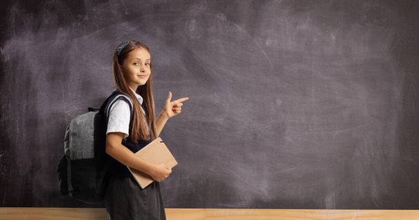 Schoolgirl with a backpack and book pointing at a blackboard and looking at camera - Фото, зображення