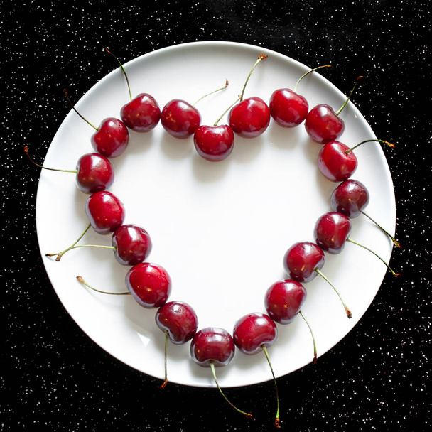 Ripe red cherries on a heart-shaped plate with an empty white plate in the center. Sweet red seasonal fruits. Flat lay. Black and white background - Photo, image