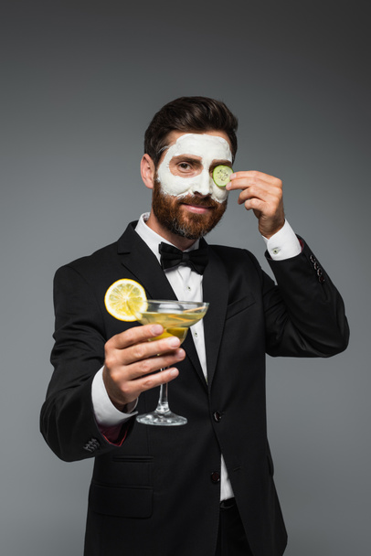 happy man in suit with clay mask on face holding cocktail isolated on grey  - Foto, Bild