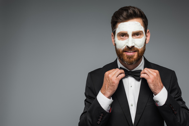 happy and bearded man in suit with clay mask on face adjusting bow tie isolated on grey - Fotoğraf, Görsel
